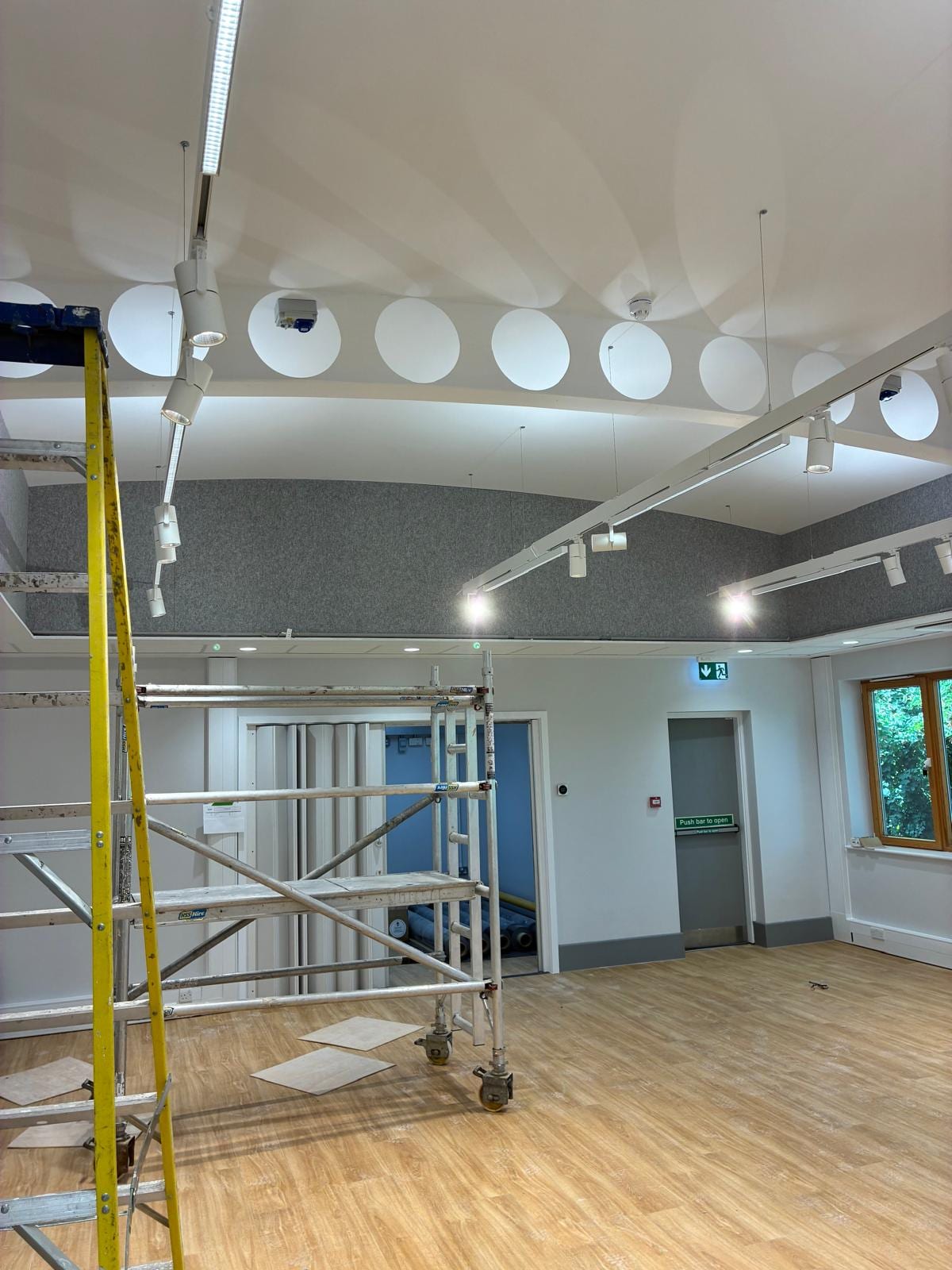 Hall with grey acoustic ceiling rafts, circular acoustic discs and track lighting, with a mobile tower still in place